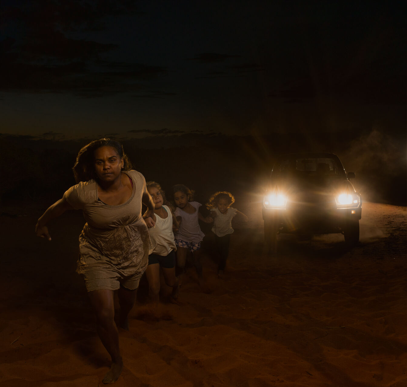A mother and children running on red sand as headlights approach